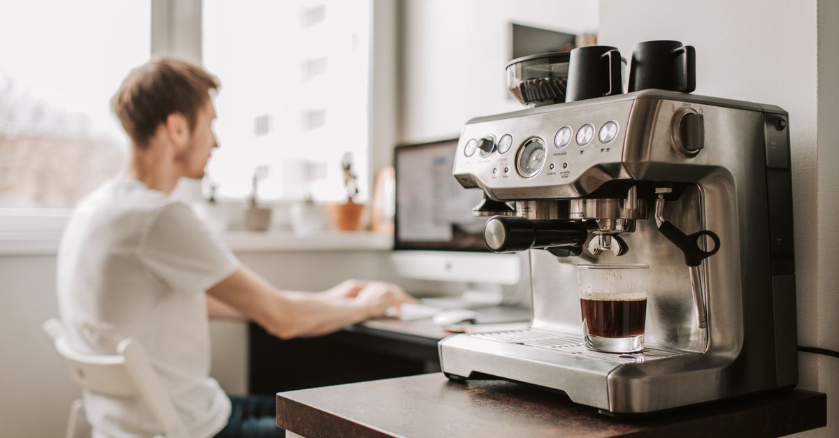 Side view of male remote worker in t shirt typing on desktop computer while sitting on plastic chair near window and coffeemaker in flat