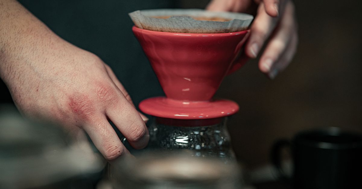 Hands preparing coffee using a red pour-over device and filter paper.